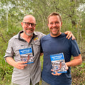 Two men standing in a forest, holding books titled 'Reptiles of Sydney'.